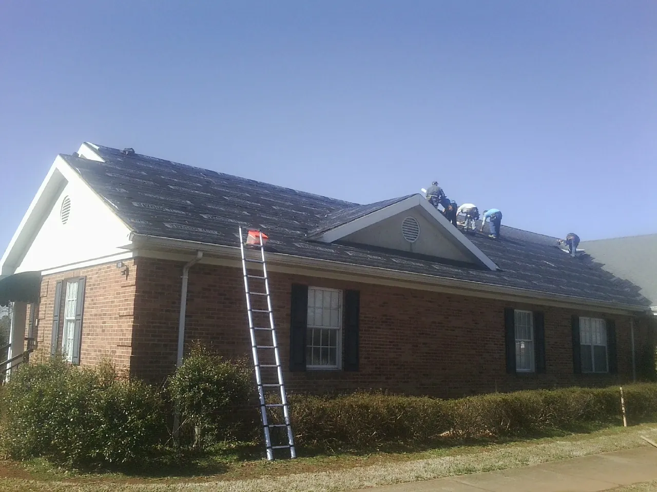 Crew tearing shingles off a brick ranch roof with a ladder set — Emergency Roof Repair work by our crew in Gilby