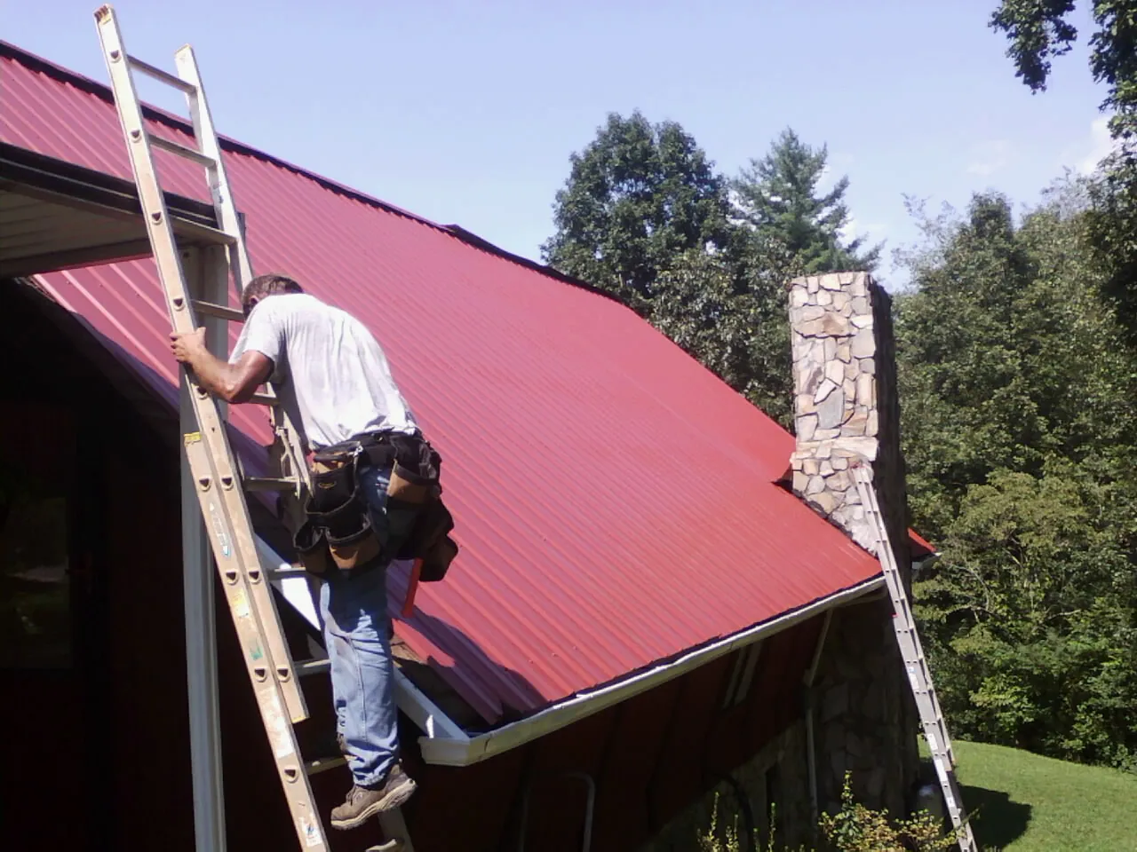 Roofer on a ladder beside a red metal roof and stone chimney — Soffit & Fascia Repair by Roofing Brothers in Gilby